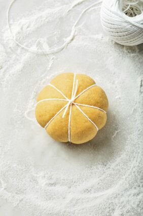 Eine Kugel mittig auf ein Stück Küchengarn legen. Mit beiden Schnurenden über der Kugel ein Kreuz bilden und das Teigstück umdrehen (wie beim Verschnüren eines Päckchens). Den Teig mithilfe des Garns in acht etwa gleich große Segmente unterteilen, dabei nicht zu fest schnüren. Zuletzt das Garn oben mittig verknoten. Auf die gleiche Weise 12 »Teigkürbisse« formen. Ein Blech mit Backpapier auslegen, die Teiglinge daraufsetzen und ca. 15 Min. an einem warmen Ort gehen lassen. Schritt 4