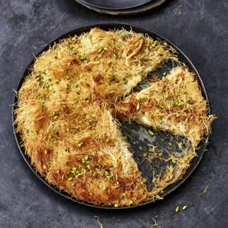 Auf dem Bild sieht man die türkische Süßspeise namens Künefe. Man sieht schön die gelblichen Fäden, die dem Dessert auch den Namen Engelshaarkuchen verleihen. Der runde Kuchen aus Kadayıf-Teig und Käse steht auf einem dunklen Hintergrund. Künefe - Engelshaarkuchen