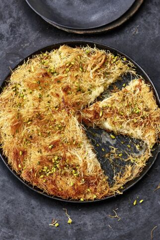 Auf dem Bild sieht man die türkische Süßspeise namens Künefe. Man sieht schön die gelblichen Fäden, die dem Dessert auch den Namen Engelshaarkuchen verleihen. Der runde Kuchen aus Kadayıf-Teig und Käse steht auf einem dunklen Hintergrund. Künefe - Engelshaarkuchen